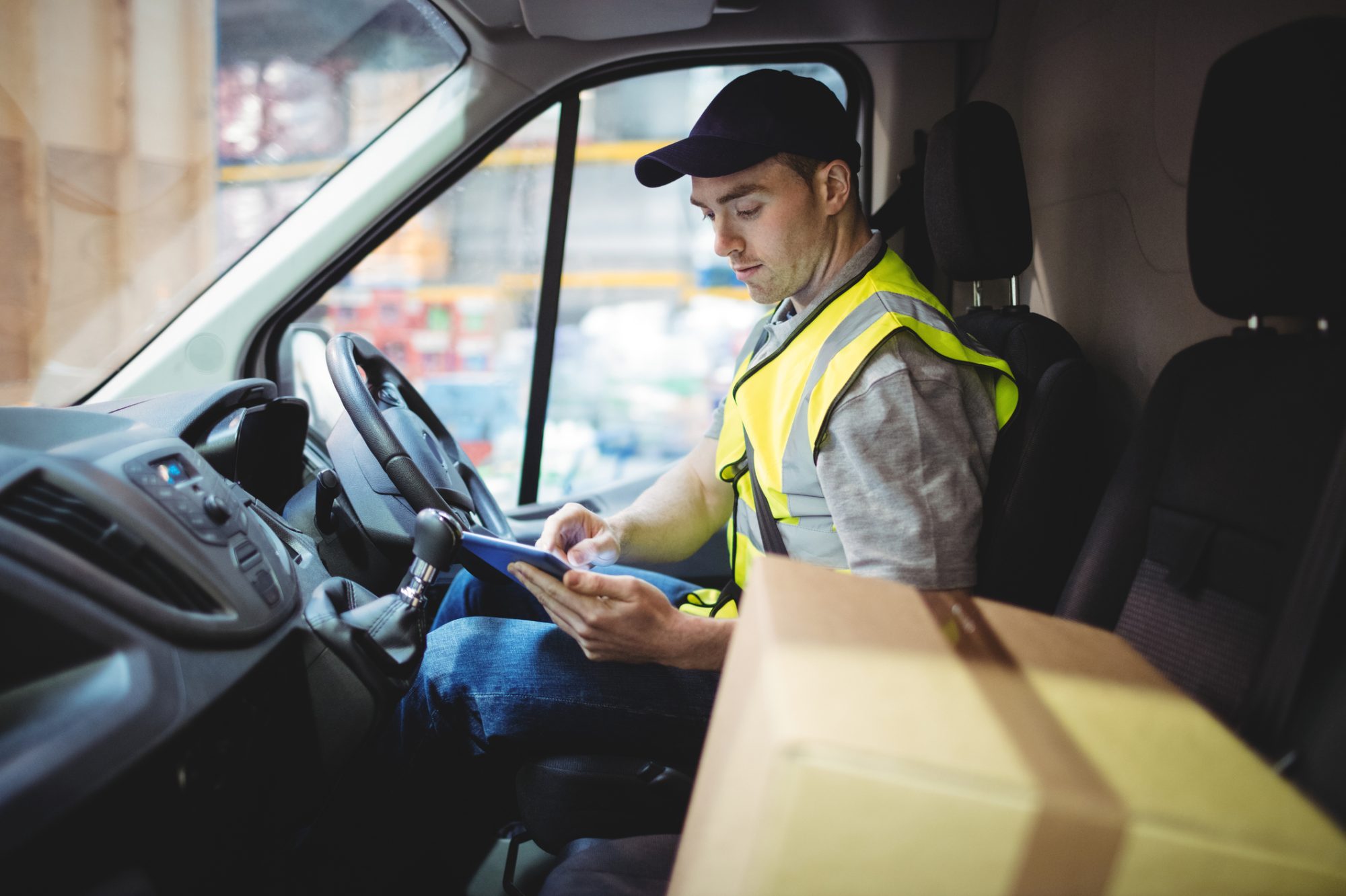 Delivery driver using tablet in van with parcels on seat outside warehouse